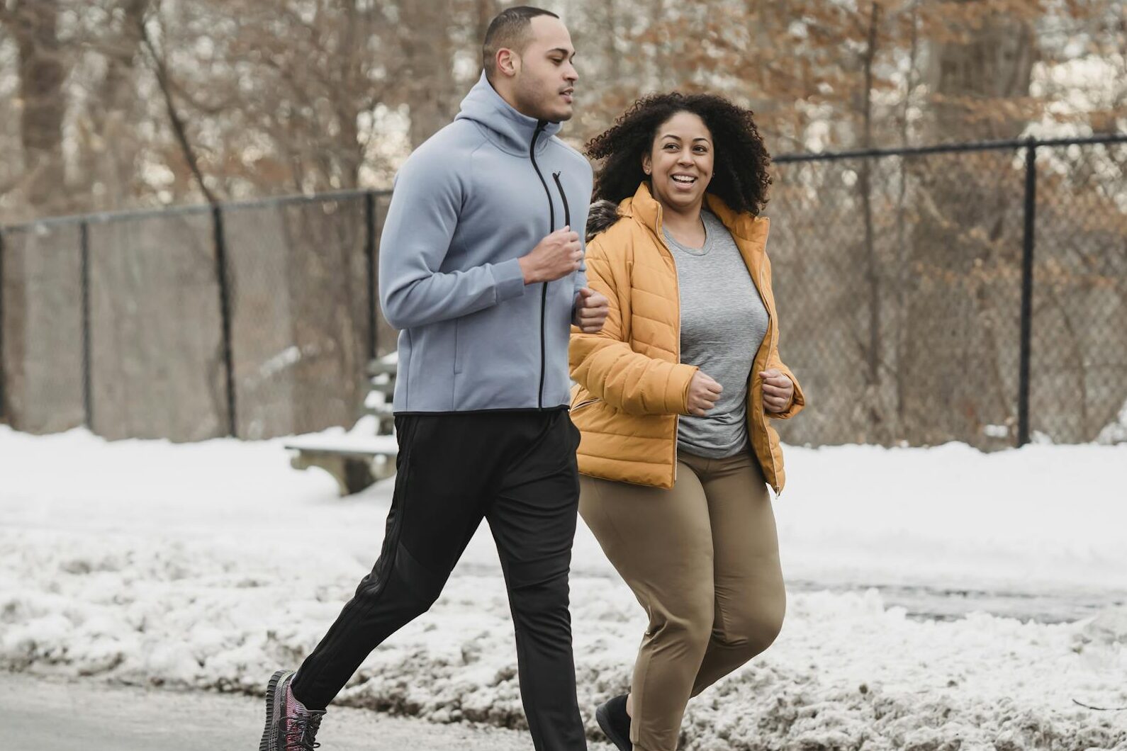 Full body content young ethnic male fitness trainer and plump black woman in warm jacket running together along snowy walkway on winter day